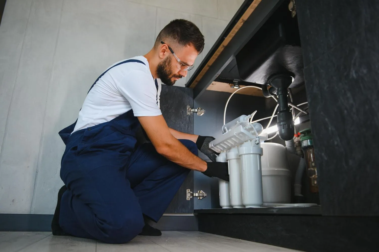 A man is kneeling down under a sink fixing a water filter.